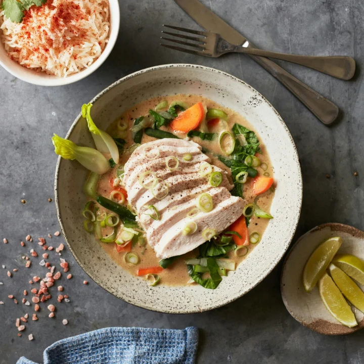 Overhead view of a bowl of sliced chicken breast in a creamy coconut broth with pak choi, carrot and spring onion, served with a bowl of rice and lime wedges