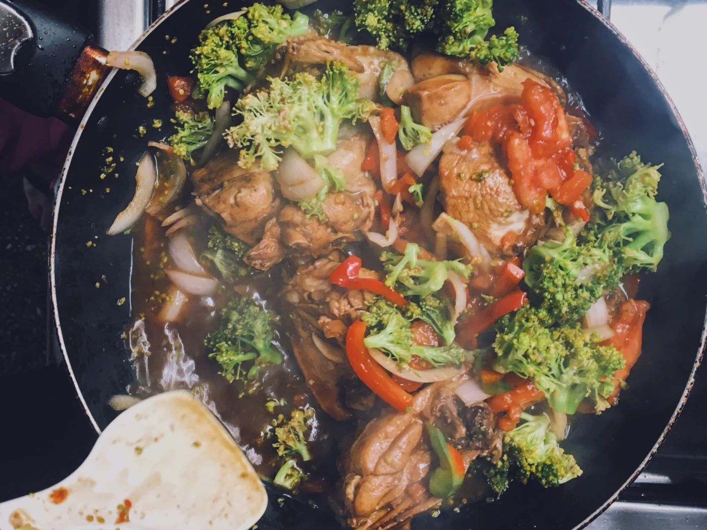 Overhead view of a frying pan on a stove holding chicken pieces in a dark sauce with broccoli florets, red pepper strips, onion and tomato, with a wooden spatula resting at the edge
