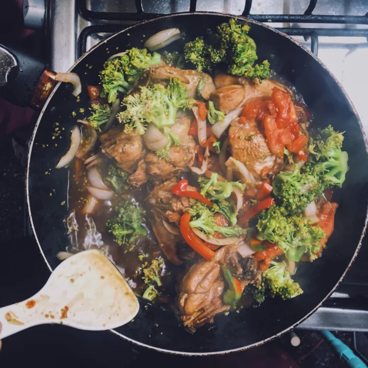 Overhead view of a frying pan on a stove holding chicken pieces in a dark sauce with broccoli florets, red pepper strips, onion and tomato, with a wooden spatula resting at the edge