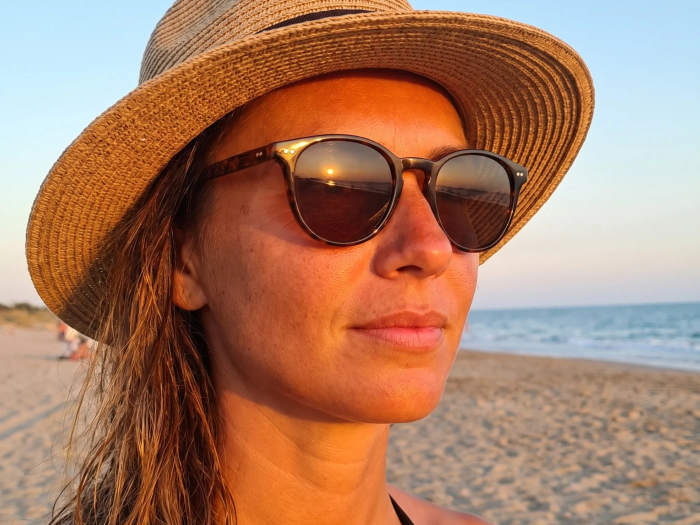 A woman wearing a straw sun hat and sunglasses on a beach at sunset, with the sea and sand behind her