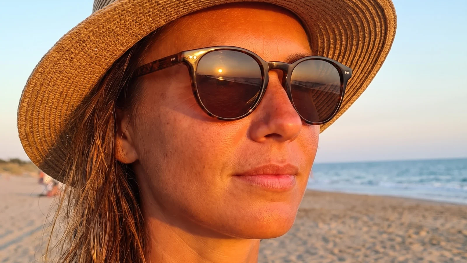 A woman wearing a straw sun hat and sunglasses on a beach at sunset, with the sea and sand behind her