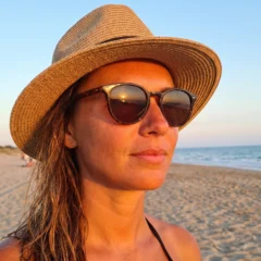 A woman wearing a straw sun hat and sunglasses on a beach at sunset, with the sea and sand behind her