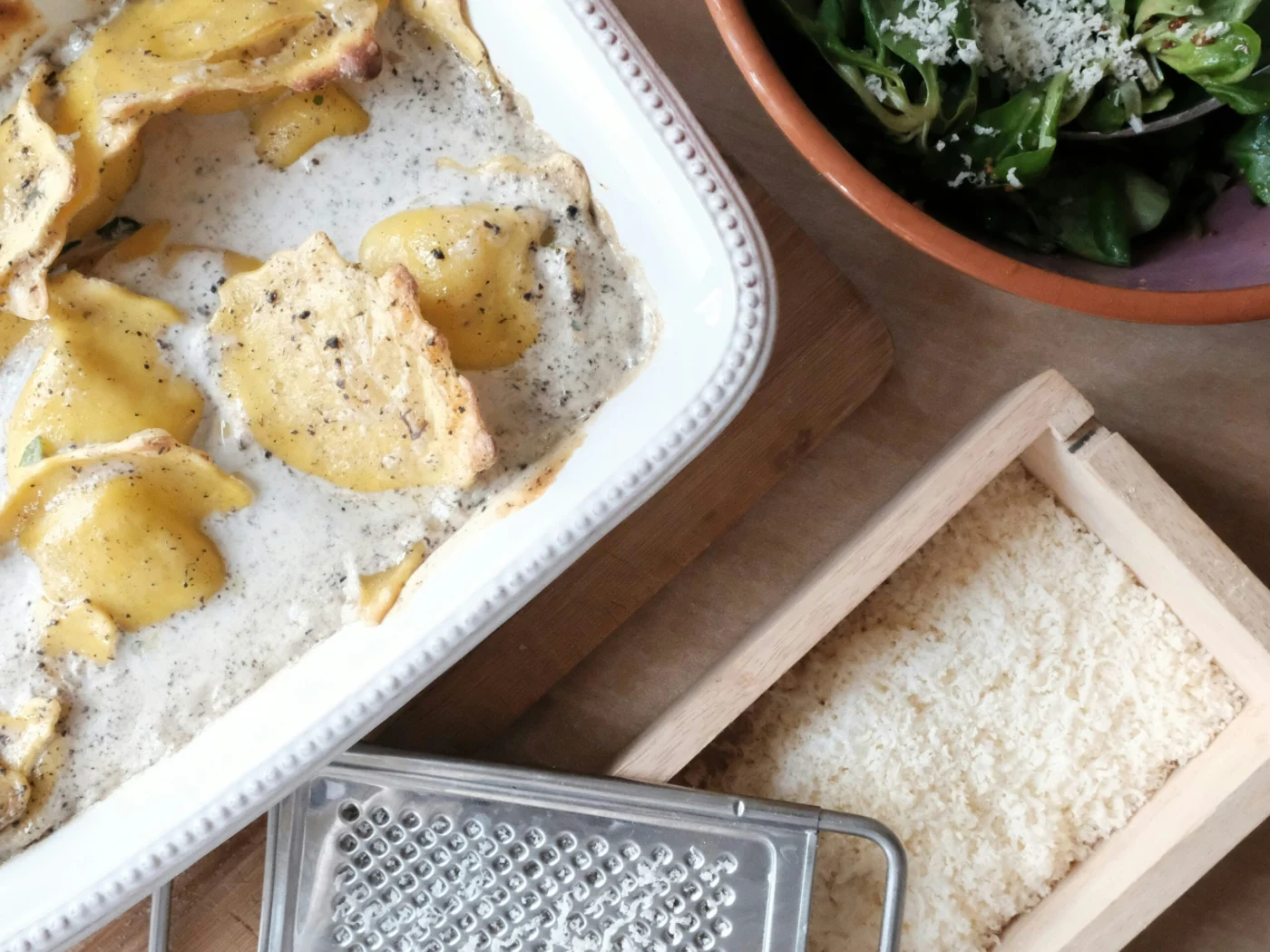 A white dish of baked ravioli in a creamy herb sauce on a wooden board, beside a bowl of green salad topped with grated cheese and a grater on a tray of grated cheese