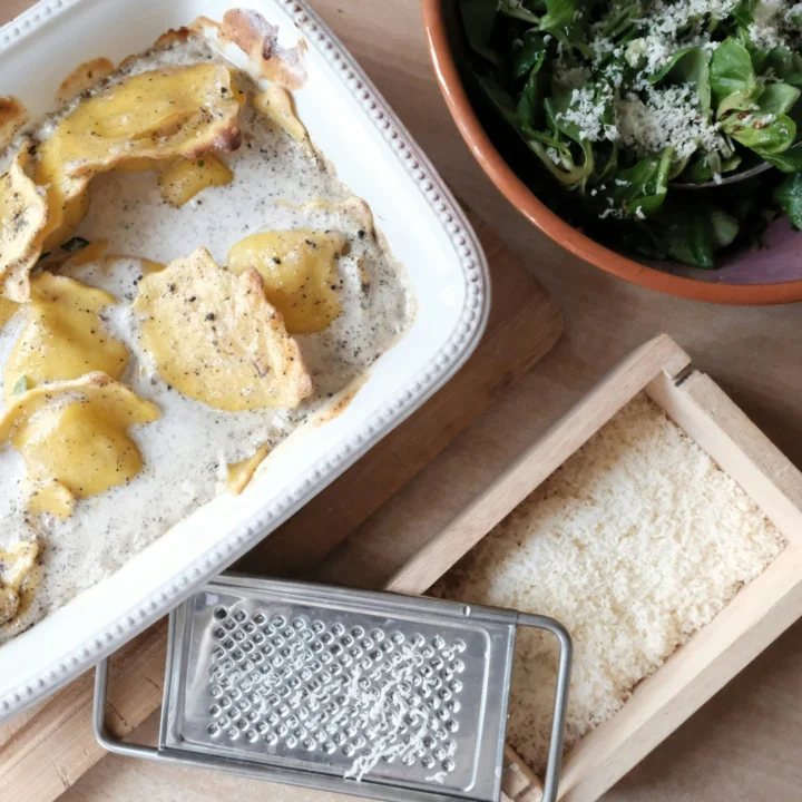 A white dish of baked ravioli in a creamy herb sauce on a wooden board, beside a bowl of green salad topped with grated cheese and a grater on a tray of grated cheese